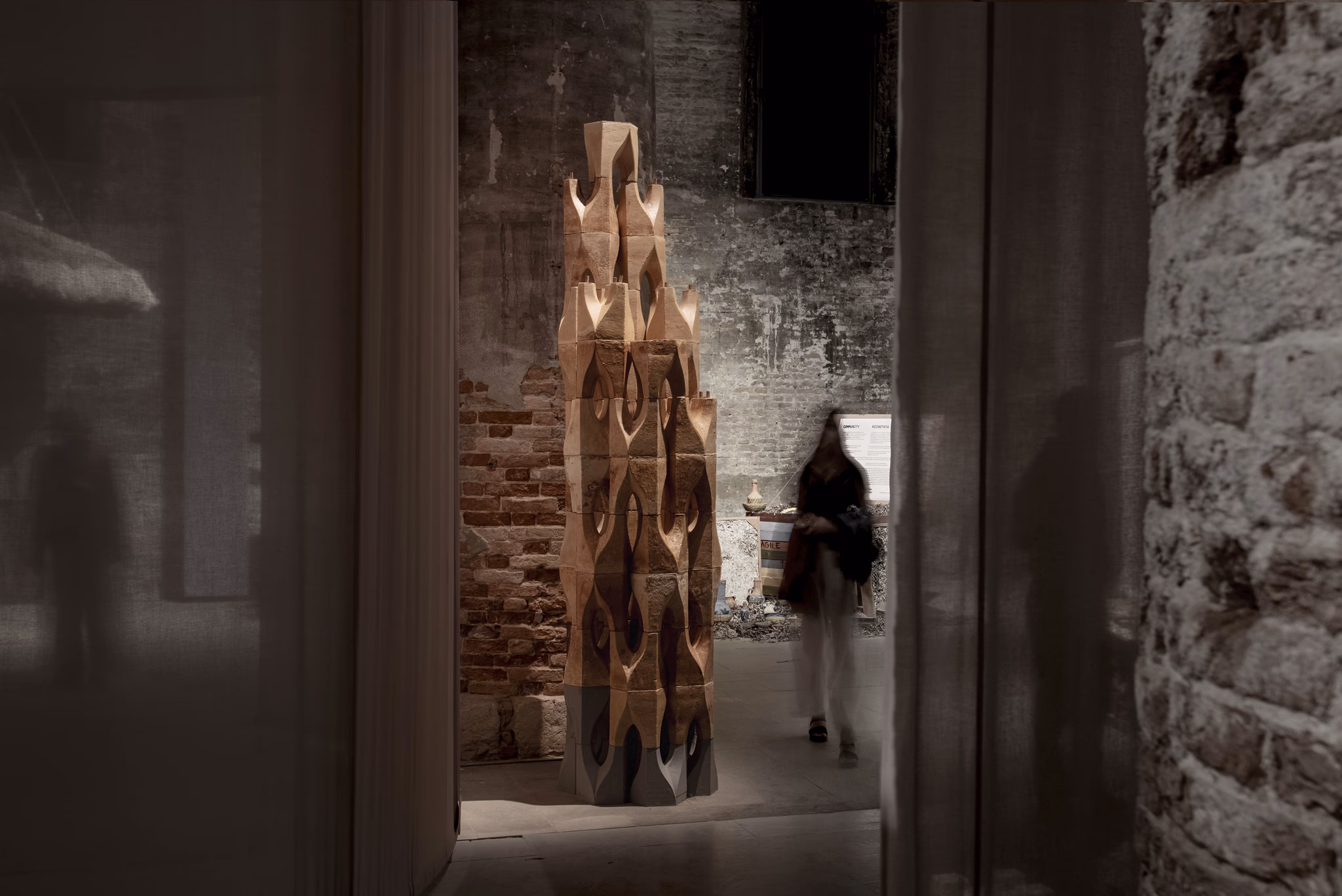A tower of mycelium blocks in a dim brick gallery, a visitor passing behind