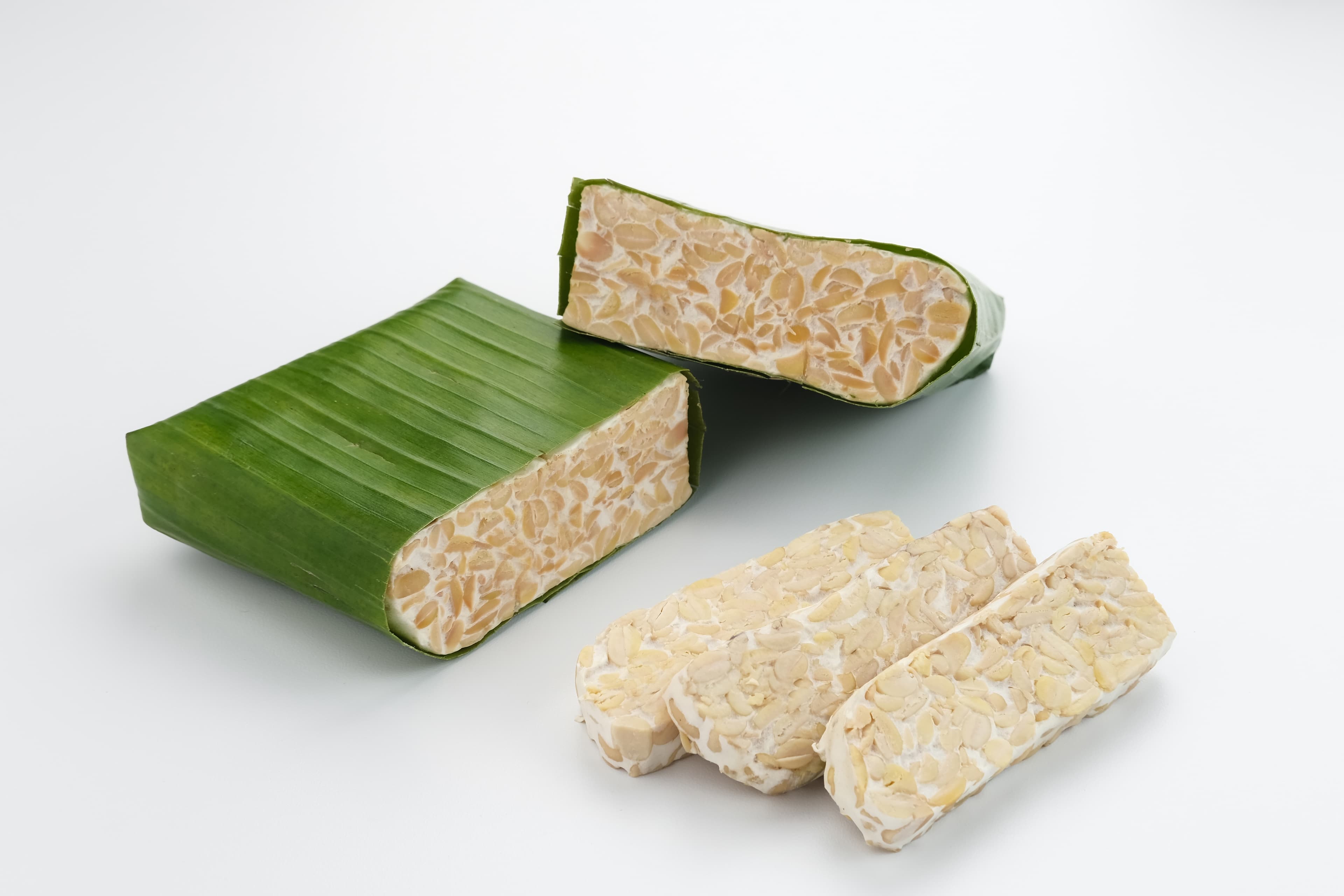Blocks of fresh tempe wrapped in banana leaf, with a few slices beside them